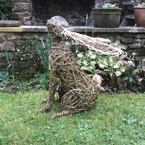 May include: A woven willow sculpture of a hare sitting on a bed of green grass. The hare has a long, flowing tail and its ears are raised.