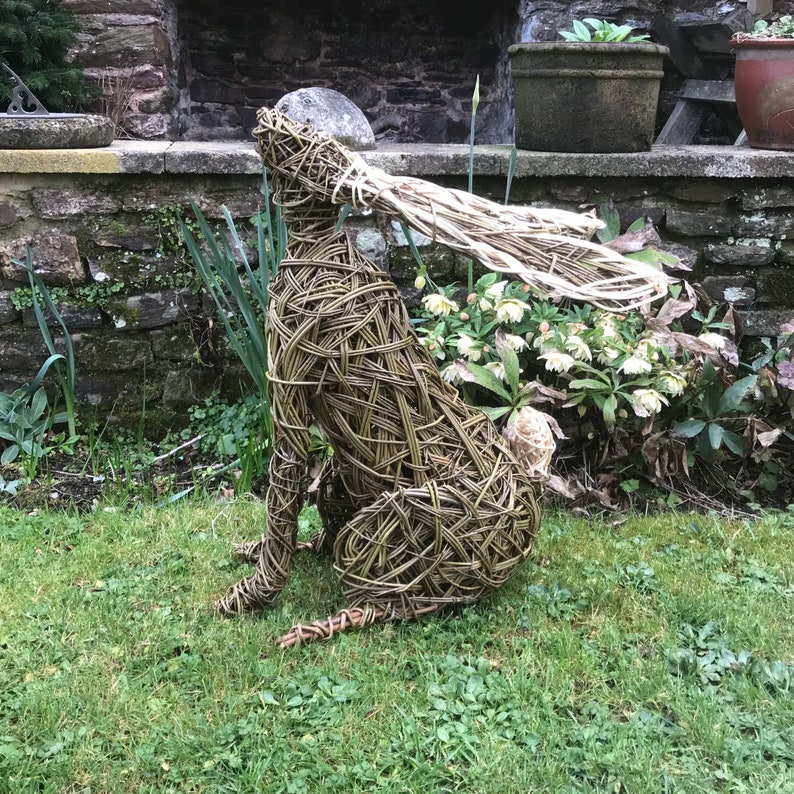 May include: A woven willow sculpture of a hare sitting on a bed of green grass. The hare has a long, flowing tail and its ears are raised.