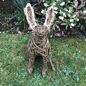 May include: A woven willow rabbit sculpture with white fabric ears. The sculpture is sitting on a bed of green grass.