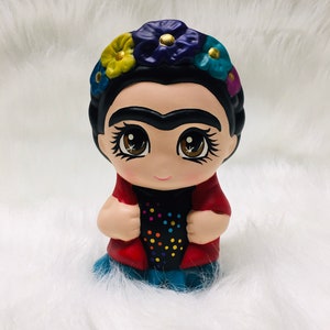 May include: A small ceramic figurine of Frida Kahlo with a colorful flower crown, red jacket, black shirt with colorful dots, and blue pants.