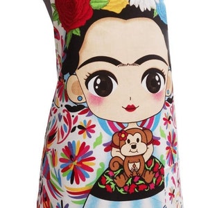 Puede incluir: Un delantal blanco con un estampado floral colorido y una imagen de dibujos animados de Frida Kahlo sosteniendo un mono. El delantal tiene una correa negra y un fondo blanco.