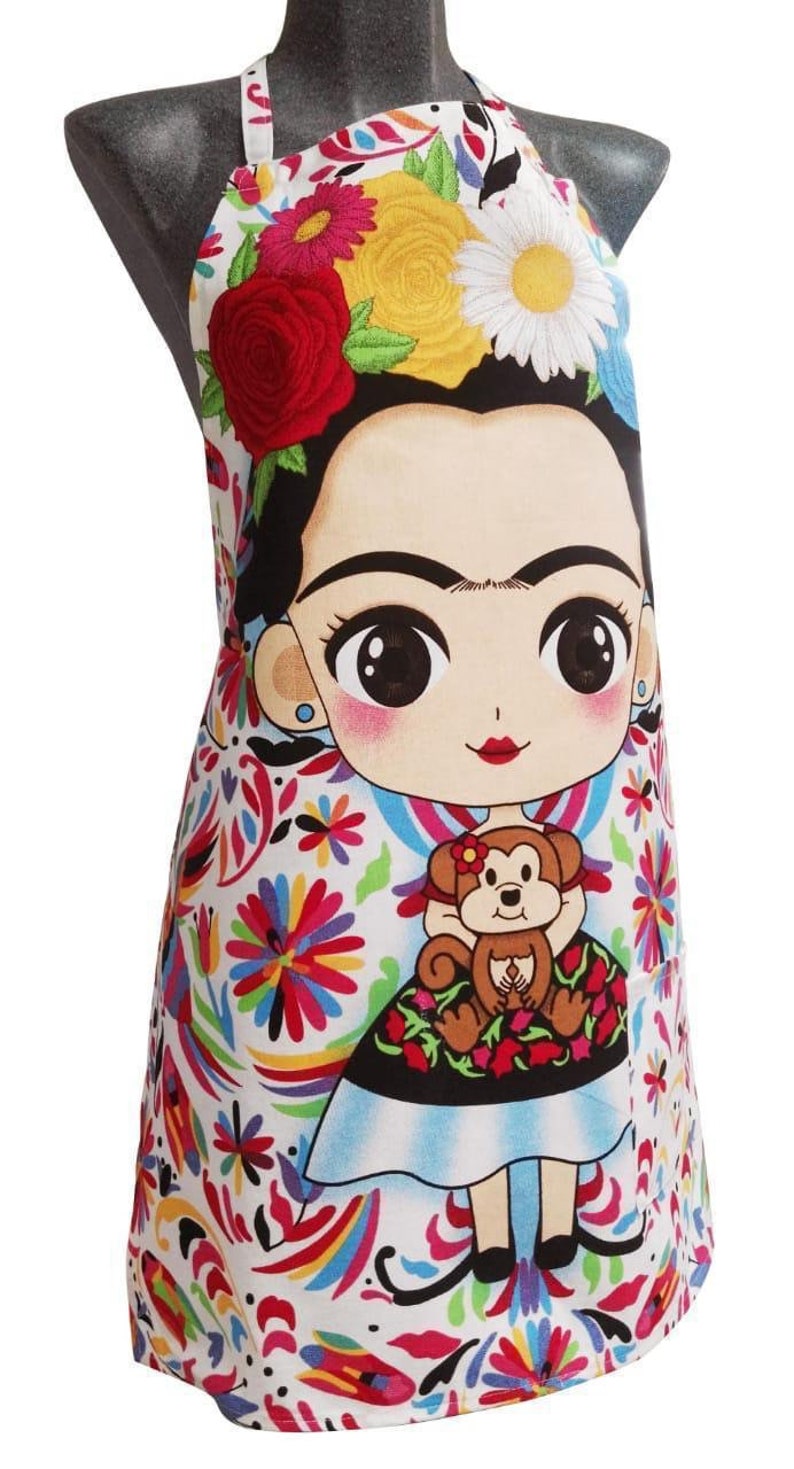 Puede incluir: Un delantal blanco con un estampado floral colorido y una imagen de dibujos animados de Frida Kahlo sosteniendo un mono. El delantal tiene una correa negra y un fondo blanco.