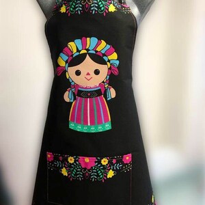 Puede incluir: Delantal negro con un dise&ntilde;o de mu&ntilde;eca mexicana colorido. La mu&ntilde;eca lleva un vestido rosa, verde y amarillo con un tocado azul y amarillo. El delantal tiene un borde floral y un bolsillo.