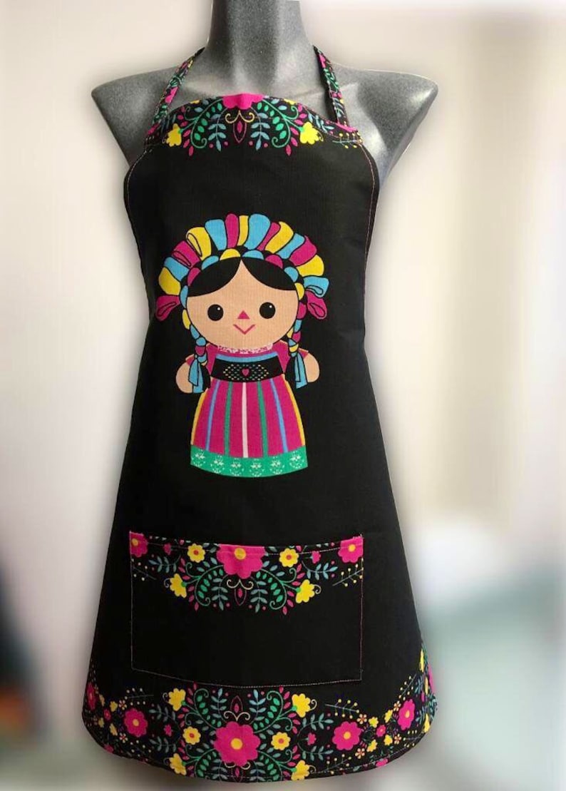 Puede incluir: Delantal negro con un dise&ntilde;o de mu&ntilde;eca mexicana colorido. La mu&ntilde;eca lleva un vestido rosa, verde y amarillo con un tocado azul y amarillo. El delantal tiene un borde floral y un bolsillo.