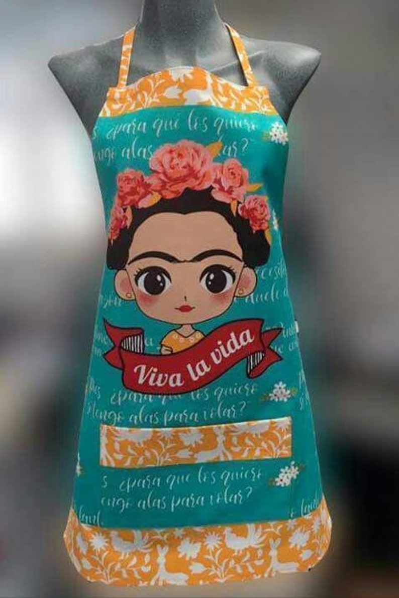 Puede incluir: Un delantal turquesa con un bolsillo presenta una ilustraci&oacute;n colorida de Frida Kahlo con el texto "Viva la vida" y texto en espa&ntilde;ol. El delantal tiene un borde con un patr&oacute;n floral amarillo y blanco.