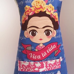 Puede incluir: Un delantal azul con una ilustraci&oacute;n colorida de Frida Kahlo. El delantal tiene un bolsillo grande con el texto "Viva la vida" y la frase "&iquest;para qu&eacute; los quiero, engo alas para volar?" en espa&ntilde;ol.
