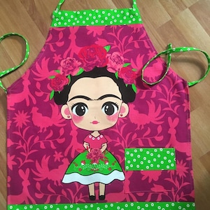 Puede incluir: Un delantal rosa con un borde verde y un bolsillo. El delantal presenta una imagen de dibujos animados de Frida Kahlo con flores en el pelo y un ramo de flores en las manos.