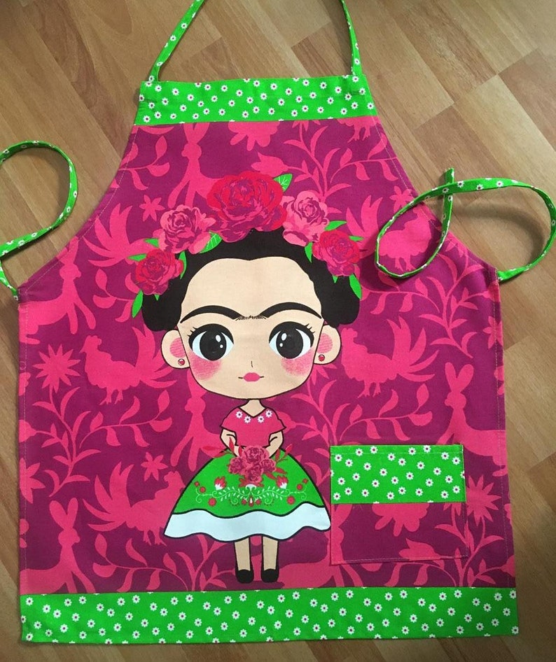 Puede incluir: Un delantal rosa con un borde verde y un bolsillo. El delantal presenta una imagen de dibujos animados de Frida Kahlo con flores en el pelo y un ramo de flores en las manos.