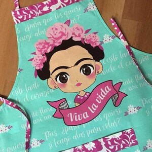 Puede incluir: Un delantal turquesa con un estampado floral rosa y una imagen de dibujos animados de Frida Kahlo. El delantal tiene una gran cinta rosa con el texto "Viva la vida" en blanco. El delantal tiene un bolsillo rosa con un estampado floral.