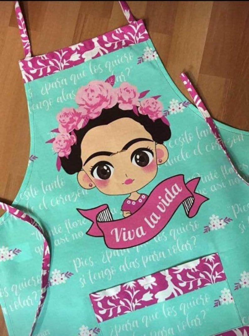 Puede incluir: Un delantal turquesa con un estampado floral rosa y una imagen de dibujos animados de Frida Kahlo. El delantal tiene una gran cinta rosa con el texto "Viva la vida" en blanco. El delantal tiene un bolsillo rosa con un estampado floral.