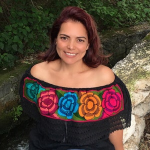 May include: A woman wearing a black off-the-shoulder top with colorful embroidered flowers. The top has a lace trim and the flowers are red, orange, blue, and green.