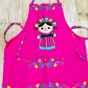 Puede incluir: Un delantal de cocina rosa con un dise&ntilde;o de mu&ntilde;eca bordada de colores. El delantal tiene un bolsillo y un ribete floral.