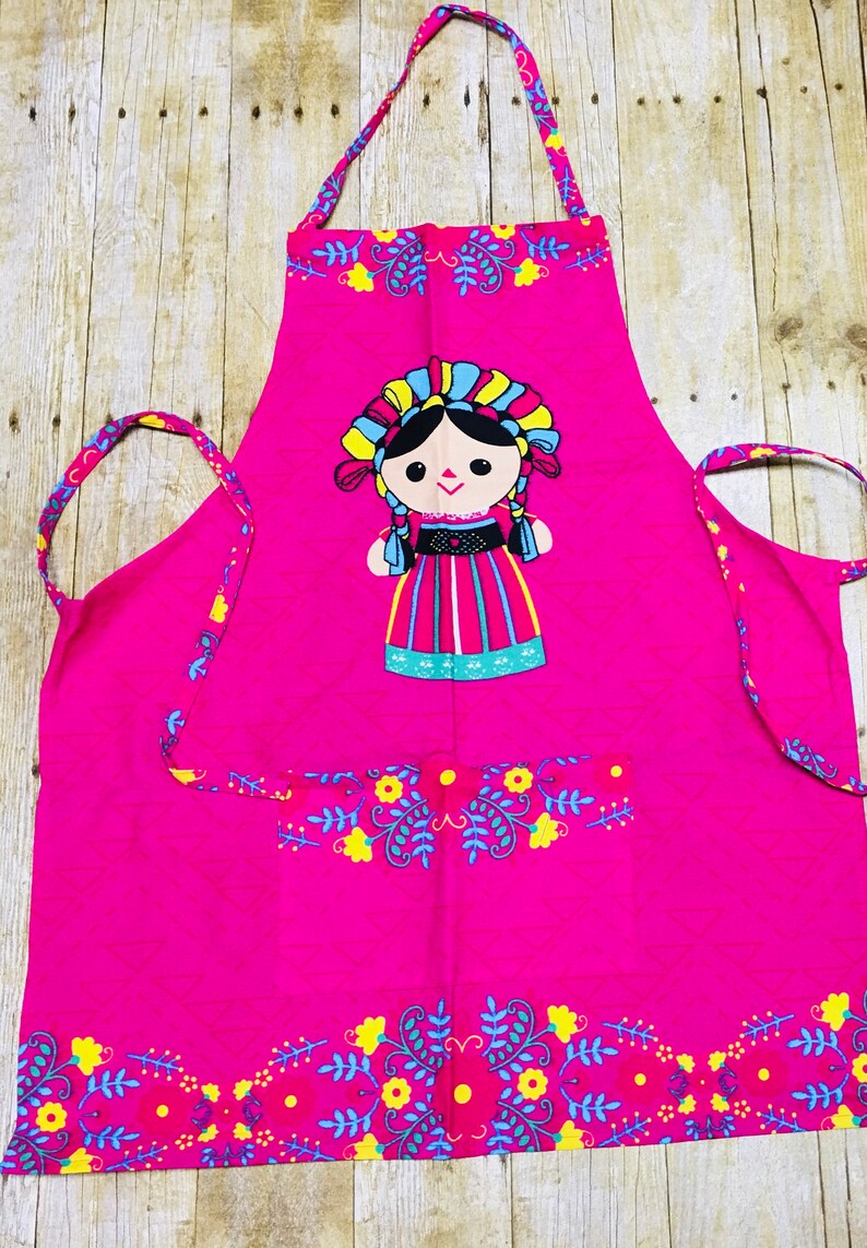 Puede incluir: Un delantal de cocina rosa con un dise&ntilde;o de mu&ntilde;eca bordada de colores. El delantal tiene un bolsillo y un ribete floral.