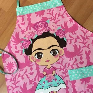 Puede incluir: Delantal rosa y verde con un patr&oacute;n floral y una imagen de dibujos animados de Frida Kahlo. El delantal tiene un bolsillo en el lado derecho.