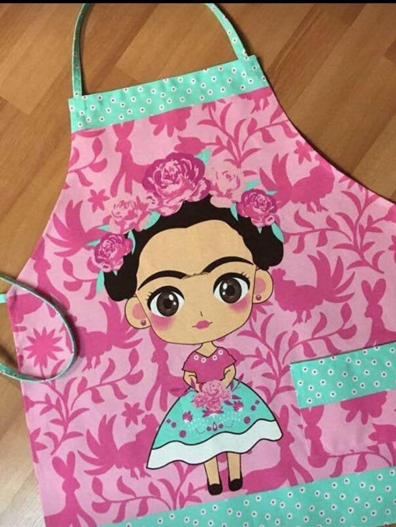 Puede incluir: Delantal rosa y verde con un patr&oacute;n floral y una imagen de dibujos animados de Frida Kahlo. El delantal tiene un bolsillo en el lado derecho.