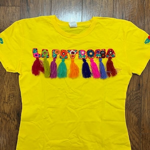May include: Yellow t-shirt with the word "LA PATRONA" embroidered in colorful letters with tassels.