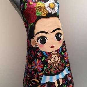 Puede incluir: Delantal negro con un estampado floral colorido y una imagen de dibujos animados de Frida Kahlo sosteniendo un mono. El delantal tiene un bolsillo en el lado izquierdo.