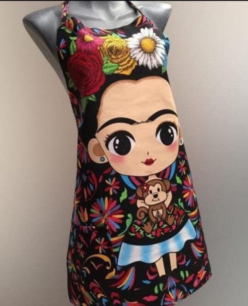 Puede incluir: Delantal negro con un estampado floral colorido y una imagen de dibujos animados de Frida Kahlo sosteniendo un mono. El delantal tiene un bolsillo en el lado izquierdo.