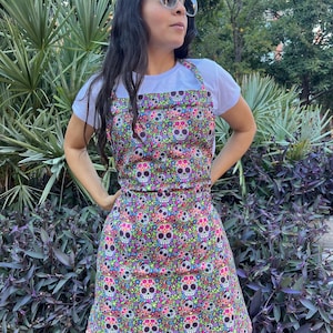 May include: A black apron with a colorful Day of the Dead skull pattern. The apron has adjustable straps and a large pocket in the front.