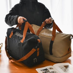 Peut inclure: Deux sacs de sport personnalisés. L'un est noir avec des sangles marron et le nom "Mahault" brodé en blanc. L'autre est beige avec des sangles marron et le nom "Jessica" brodé en blanc. Les deux sacs ont un compartiment principal zippé.