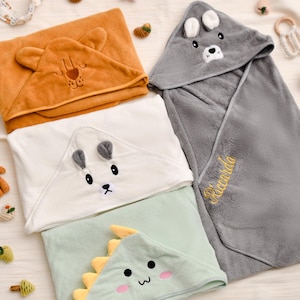 Puede incluir: Una colección de toallas con capucha para bebés en varios colores. Las toallas presentan capuchas con temas de animales, como un oso, un lobo y un dinosaurio. Una toalla gris está bordada con el nombre "Ricardo" en hilo dorado. Las toallas son suaves.