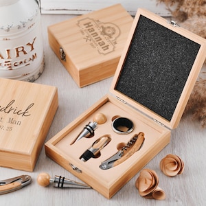 Puede incluir: Un juego de abridor de vino de madera con sacacorchos, tapón y vertedor, presentado en una caja de madera. La caja está abierta, revelando las herramientas en un inserto de espuma. También hay cajas de madera adicionales y flores decorativas.