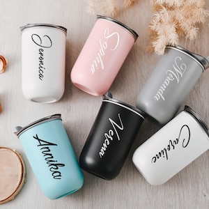 Puede incluir: Seis vasos de acero inoxidable personalizados con tapas. Los vasos son de varios colores, incluyendo blanco, rosa, gris, negro y azul. Cada vaso tiene un nombre escrito en él en letra negra: Veronica, Sophia, Miranda, Selena, Annika y Caroline.