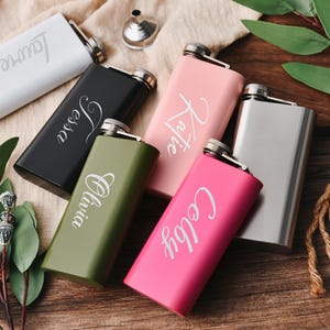 Könnte beinhalten: Fünf personalisierte Edelstahl-Taschenflaschen in verschiedenen Farben. Die Flaschen sind mit den Namen "Lauren", "Jess", "Olivia", "Katie" und "Colby" graviert.