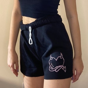 May include: Black athletic shorts with a white drawstring and a pink rhinestone heart with devil horns design.