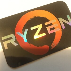 May include: A black sticker with a holographic silver and rainbow colored "RYZEN" logo. The logo is centered on a red and orange circle with a brushstroke design.