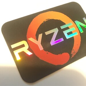 May include: A black sticker with a holographic, rainbow-colored "RYZEN" logo. The logo is surrounded by a red circle with a brushstroke design.