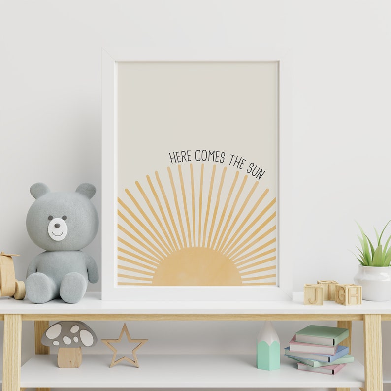 May include: A white framed print with a yellow sun graphic and the text "Here comes the sun". The print is hanging on a white wall above a wooden shelf with a gray teddy bear, a mushroom, a star, and books.
