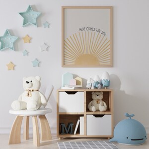 May include: A white wooden shelf unit with four cubbies, two of which are open. The top two cubbies have white drawers. The bottom two cubbies are open and contain a book and a wooden letter M. The shelf unit is decorated with a white teddy bear, a blue whale, and a variety of small toys. The wall behind the shelf unit is decorated with a framed print that reads "Here comes the sun" and several blue and yellow star-shaped decorations.