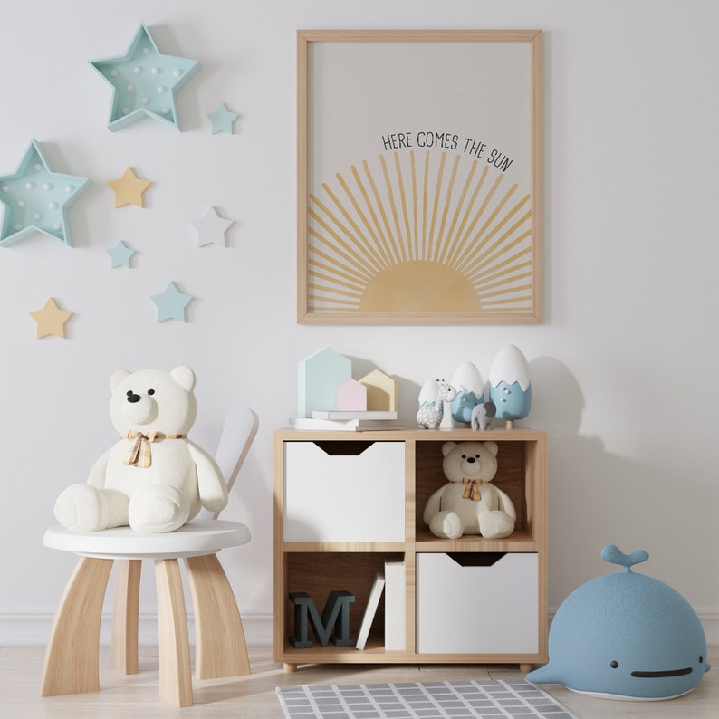 May include: A white wooden shelf unit with four cubbies, two of which are open. The top two cubbies have white drawers. The bottom two cubbies are open and contain a book and a wooden letter M. The shelf unit is decorated with a white teddy bear, a blue whale, and a variety of small toys. The wall behind the shelf unit is decorated with a framed print that reads "Here comes the sun" and several blue and yellow star-shaped decorations.