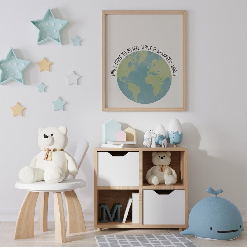 May include: A white wooden shelf unit with four cubbies, two of which are open and two of which have white drawers. The top shelf has a variety of small toys, including a white teddy bear, a blue whale, and a set of colorful wooden houses. The bottom shelf has a book with the letter "M" on the cover. The wall behind the shelf is white and decorated with blue and yellow star-shaped decorations. A framed print with the text "And I think to myself what a wonderful world" is hanging on the wall above the shelf.