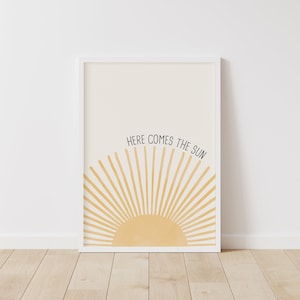 May include: A white framed print with a yellow sun rising over a white background. The text "HERE COMES THE SUN" is written in black above the sun.