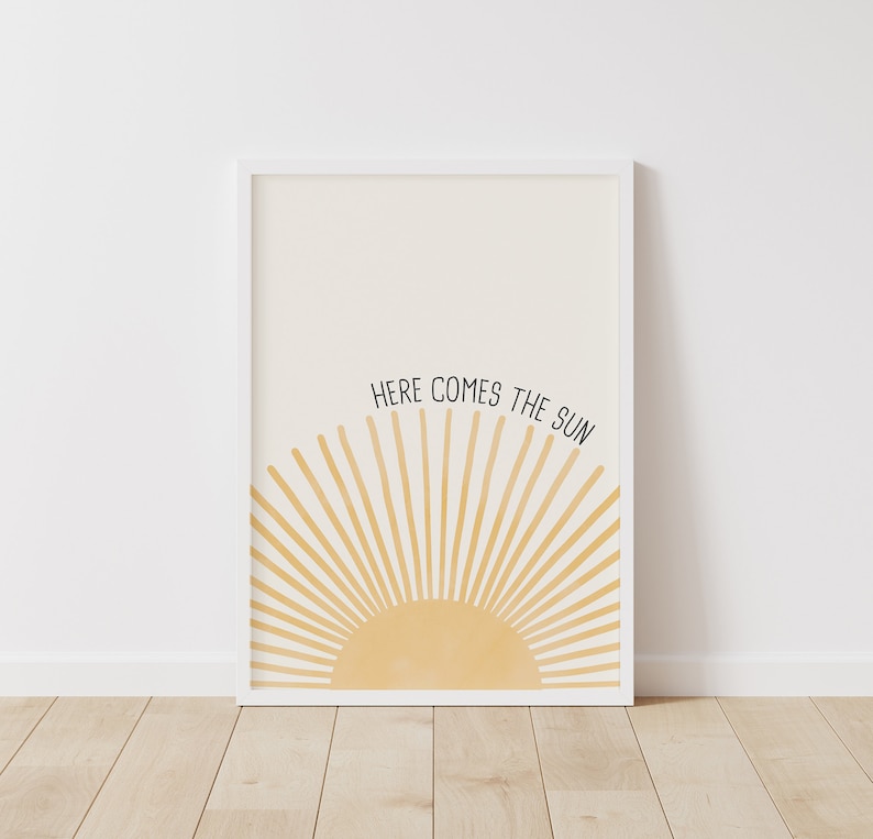 May include: A white framed print with a yellow sun rising over a white background. The text "HERE COMES THE SUN" is written in black above the sun.