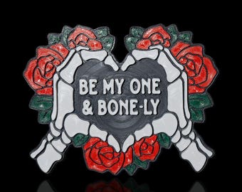 May include: A black heart-shaped pin with white skeletal hands holding a bouquet of red roses. The text "BE MY ONE & BONE-LY" is written on the heart.
