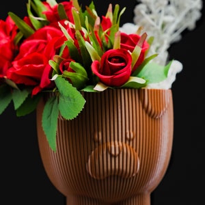 May include: A bouquet of red roses with green leaves in a brown ceramic planter shaped like a bear with a smiling face.