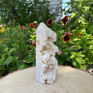 May include: A white crystal cluster with a rough, textured surface. The crystal is shaped like a tall, narrow pyramid and has a pointed top. It is sitting on a wooden surface.