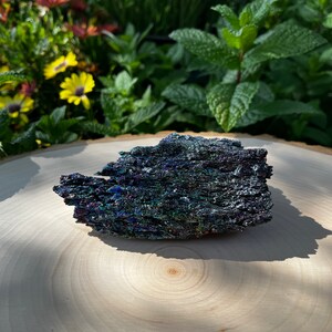 May include: A raw, iridescent silicon carbide crystal cluster featuring a jagged, metallic texture with deep shades of blue, purple, and green that shimmer across its crystalline surface, displaying a complex, multi-faceted geometric structure.