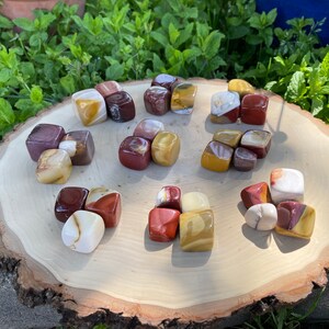 May include: A collection of 15 tumbled, polished, multi-colored jasper stones. The stones are in various shades of red, brown, yellow, and white. The stones are arranged in a circular pattern on a wooden surface.