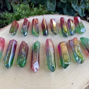 May include: A collection of 17 iridescent green, red, and gold crystal points arranged on a wooden surface. The points are all approximately the same size and shape, and they have a smooth, polished finish.