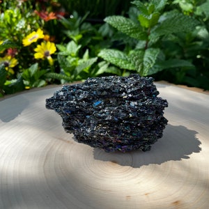 May include: A rough, jagged piece of iridescent silicon carbide crystal with a dark, metallic black base color. The surface features numerous sharp, crystalline facets that reflect light with flashes of blue, purple, and gold, creating a prismatic effect across the textured, multi-layered structure of the mineral specimen.