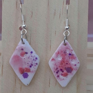 May include: A pair of diamond-shaped earrings with a white base, decorated with pink and purple accents. The earrings have a speckled design with small, irregular shapes and glitter. Silver-coloured ear wires complete the jewellery.