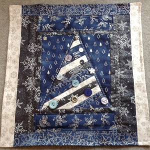 May include: A festive Christmas tree wall hanging. The tree is constructed from white and grey striped fabric and adorned with assorted coloured buttons. The background is a dark blue fabric with small white trees and snowflakes. The border is a light grey fabric with a floral pattern.
