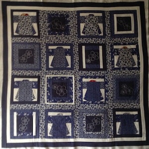 May include: A blue and white quilt with a repeating pattern of kimono-style garments and floral designs. The quilt is made up of square blocks with different patterns, including polka dots and floral prints. The quilt has a dark blue border.