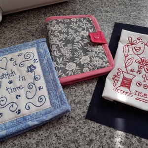 May include: Three handmade fabric items: a blue and white embroidered cover with the text "a stitch in time", a floral print notebook cover with a pink border, and a white fabric with red embroidery of sewing-related images and text.