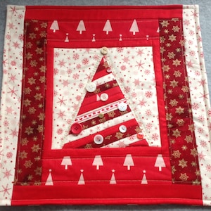 May include: A festive Christmas table runner with a quilted Christmas tree design. The tree is made of red and white striped fabric and decorated with buttons. The runner has a red border with white tree and snowflake patterns, and measures approximately 30 cm square.