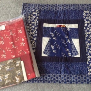 May include: A quilted placemat featuring a central kimono design in blue and white, framed by a border of blue and white floral and geometric patterns. A clear plastic bag holds red fabric with white floral patterns and other fabric pieces.